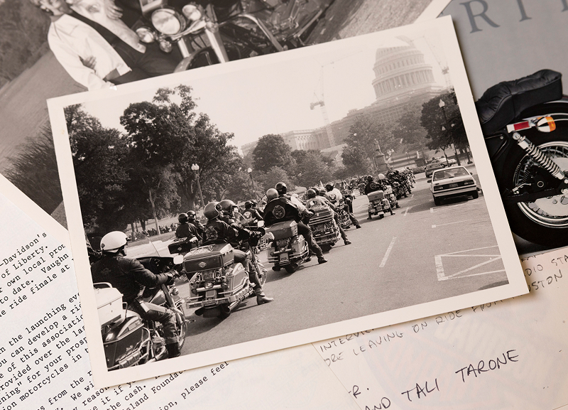 Black-and-white photo of a motorcyclist procession riding toward the U.S. Capitol, laying among papers
